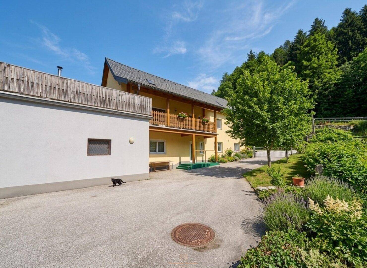 Gelbes Haus mit Holzbalkon, umgeben von viel Grün und einem gepflasterten Hof.