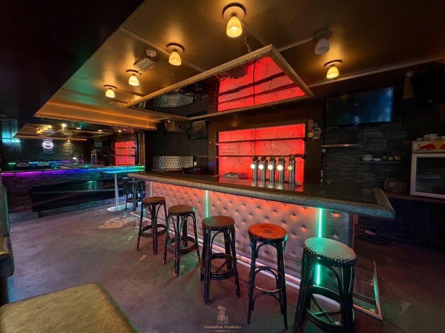 Innenansicht einer Bar mit beleuchteter Theke, Barhockern und stimmungsvoller farbiger Beleuchtung.