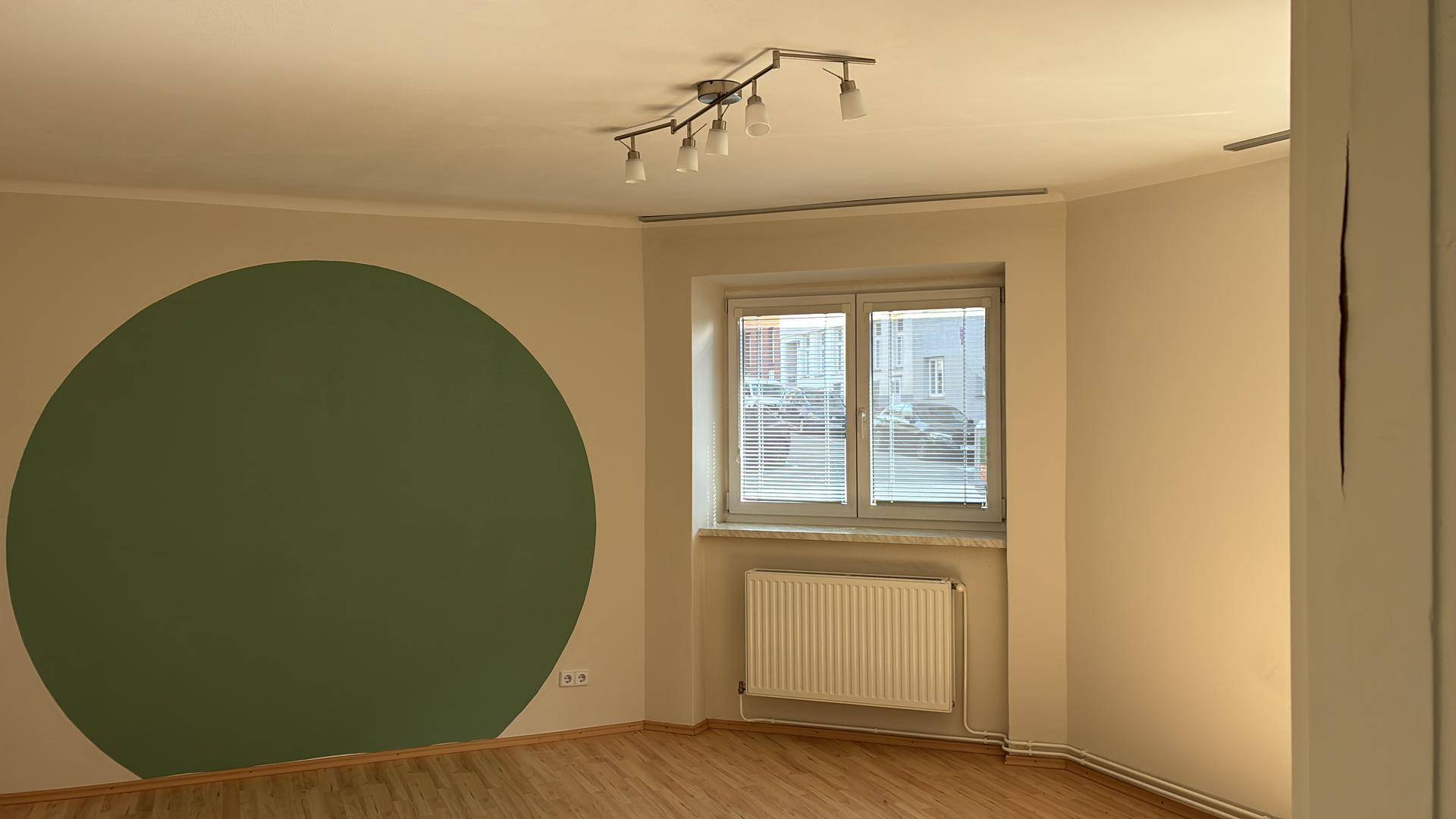 Helles Zimmer mit Laminatboden, einem großen Fenster mit Jalousien und einer grünen Kreiswand.