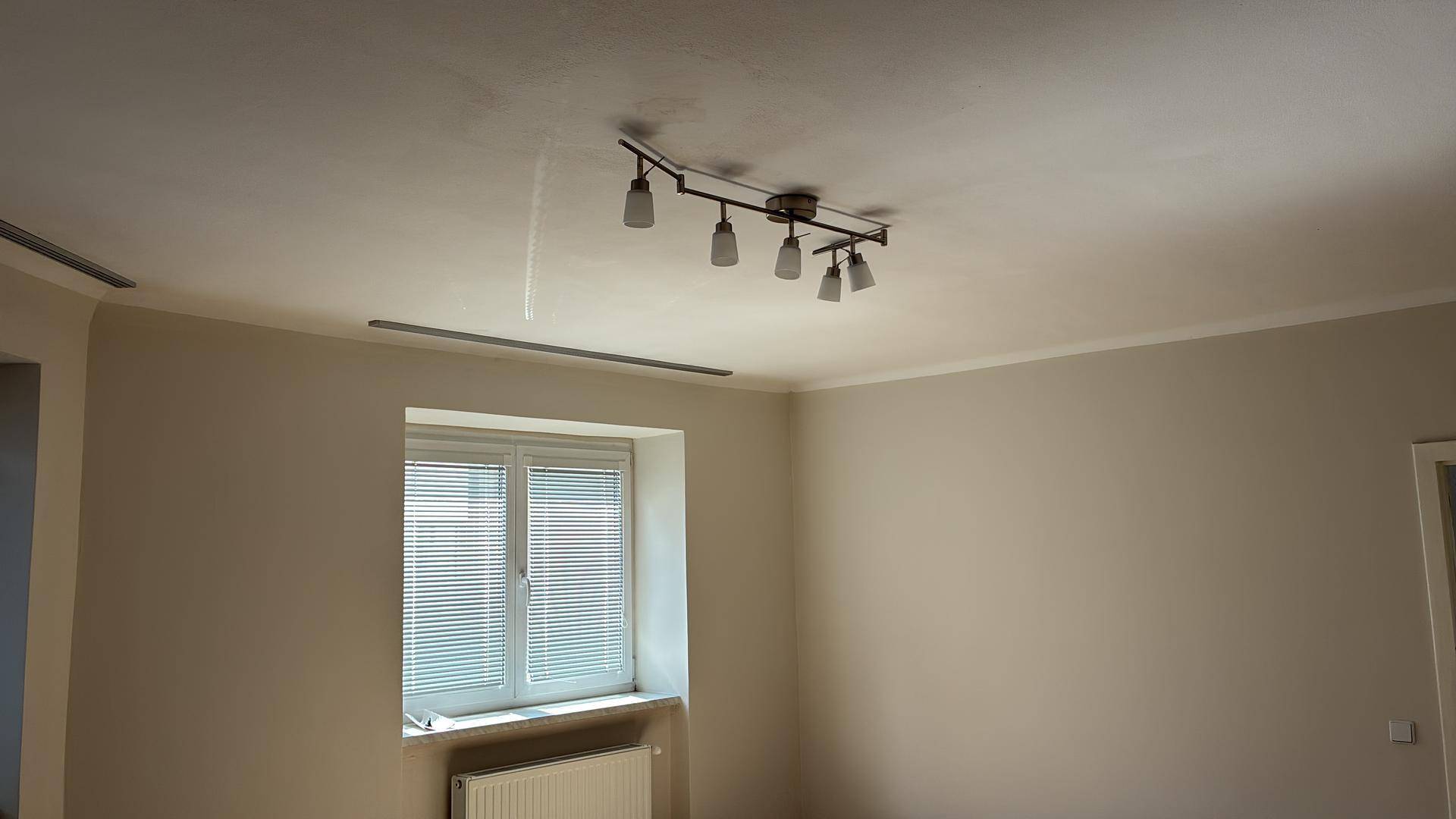 Heller Raum mit Laminatboden, einem Fenster mit Jalousien und einer modernen Deckenleuchte.