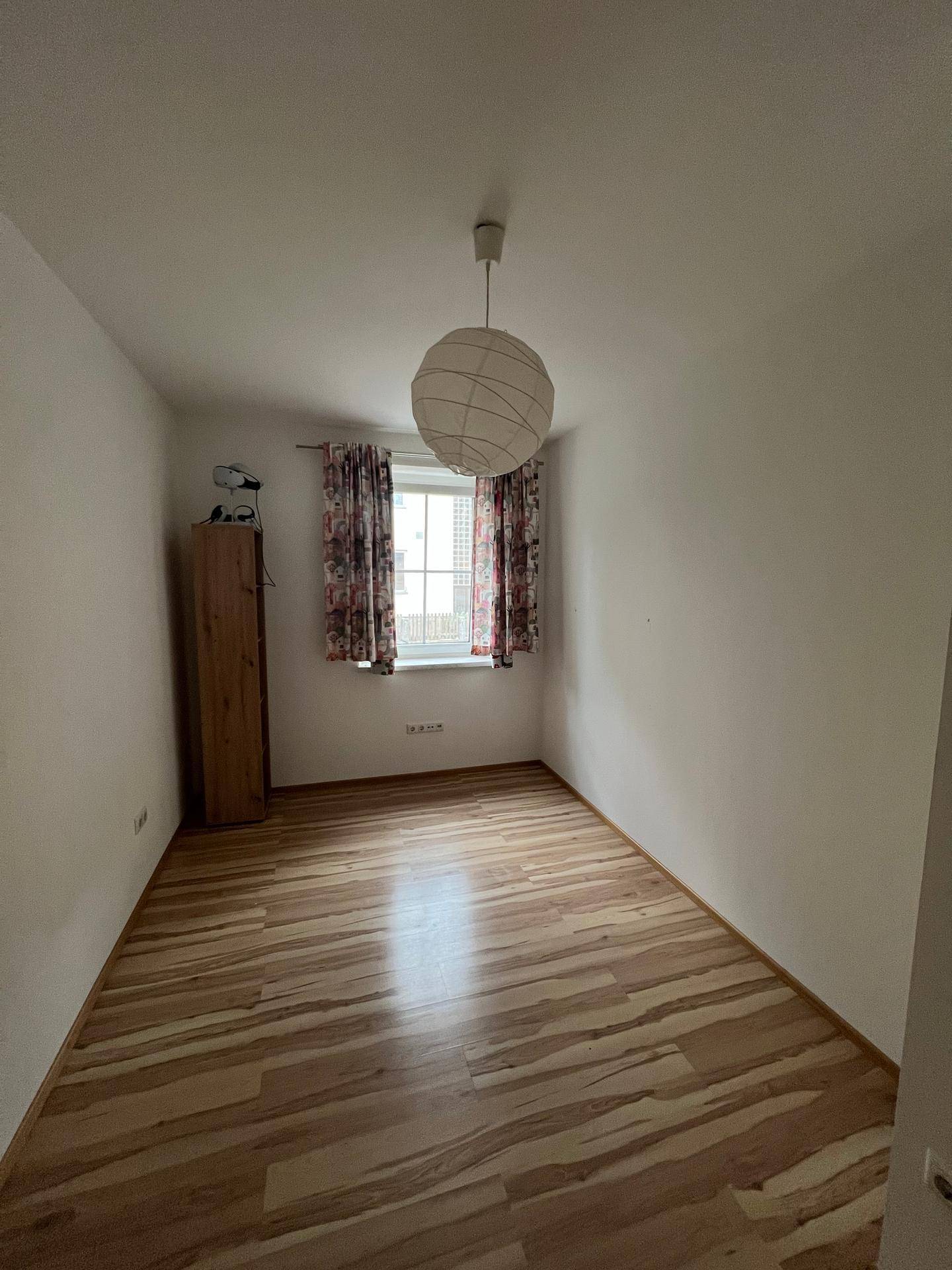 Leeres Zimmer mit Laminatboden, einem Fenster und einfacher Deckenleuchte.
