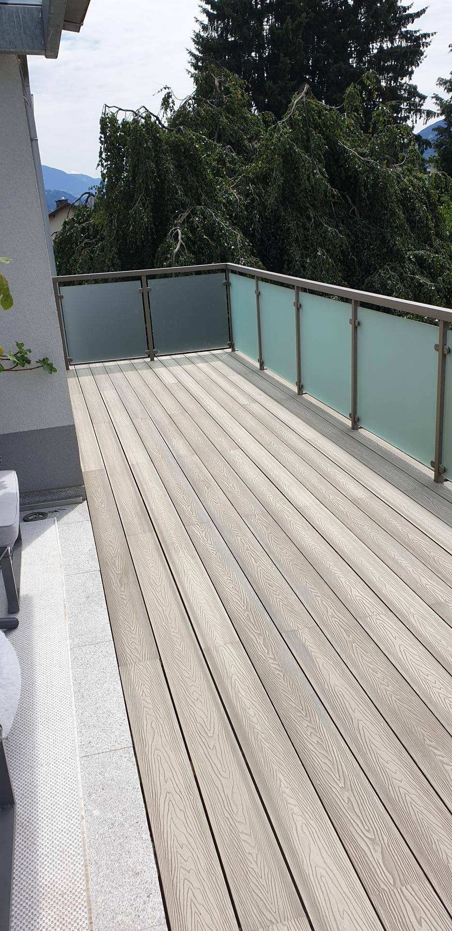 Geräumige Terrasse mit modernen Holzverbunddielen und Glasgeländer, umgeben von grünen Bäumen.