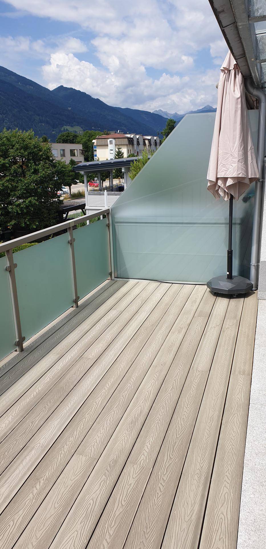 Sonniger Terrassenbereich mit Holzverbunddielen, Glasgeländer und einem Sonnenschirm, mit Bergblick.