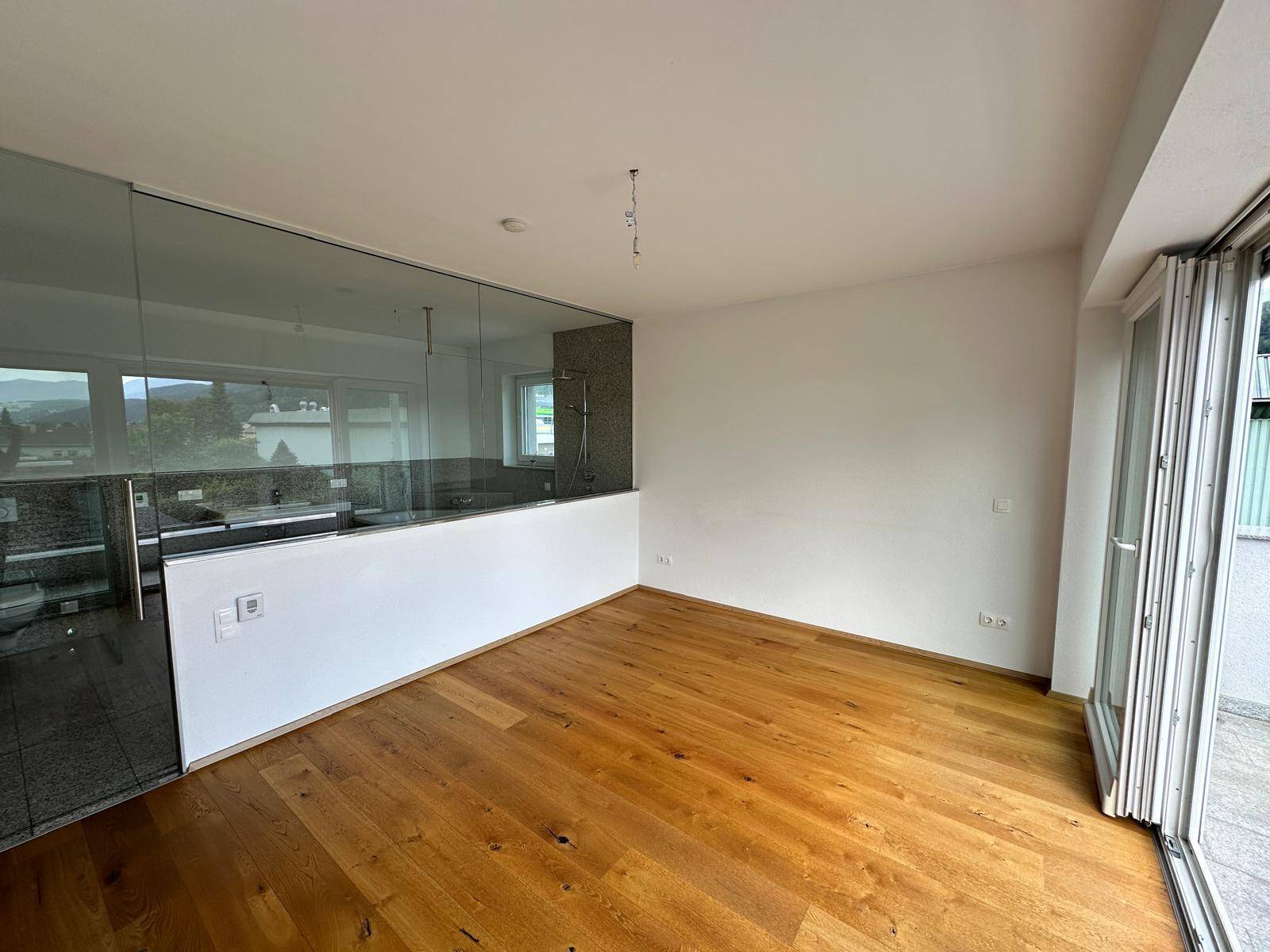 Schlafzimmer mit Holzboden und großen Fenstern, die Zugang zu einer Terrasse bieten, angrenzend an ein Badezimmer.