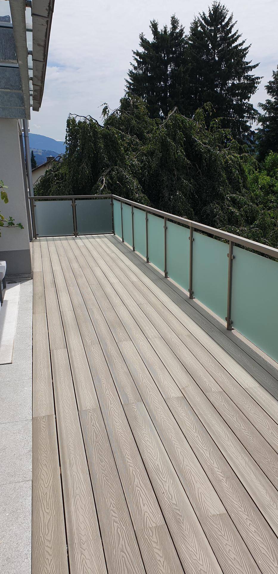 Lange Terrasse mit Holzverbunddielen und modernem Glasgeländer, umgeben von üppigem Grün.