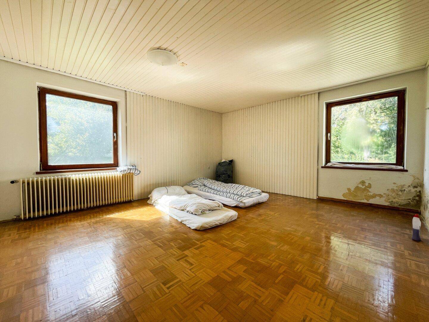 Schlafzimmer mit Parkettboden und zwei Fenstern, die viel Tageslicht hereinlassen.