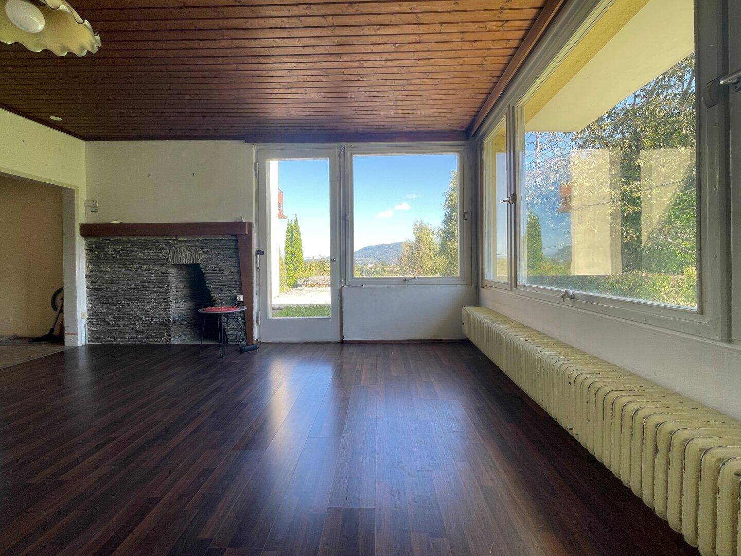 Helles Wohnzimmer mit Holzbalkendecke, dunklem Boden und Blick ins Grüne.