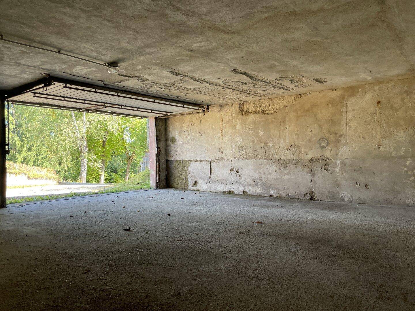 Geräumige Garage mit Betonboden und Blick ins Grüne durch das offene Tor.