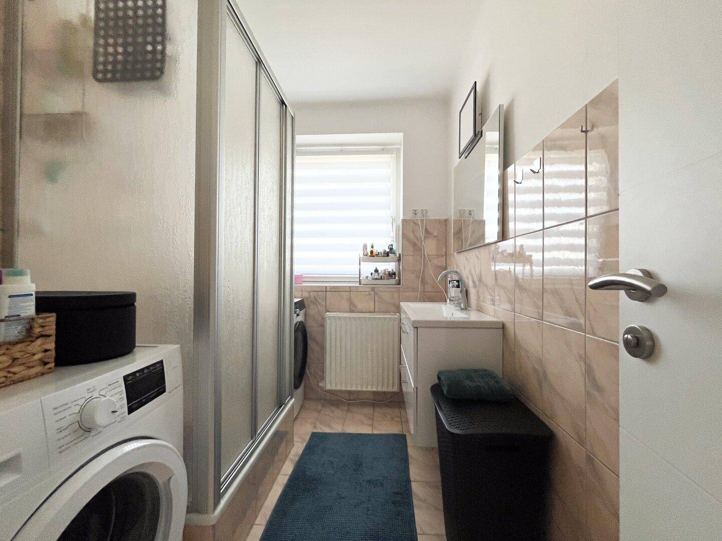 Badezimmer mit Duschkabine, Waschmaschine und hellbraunen Fliesen.