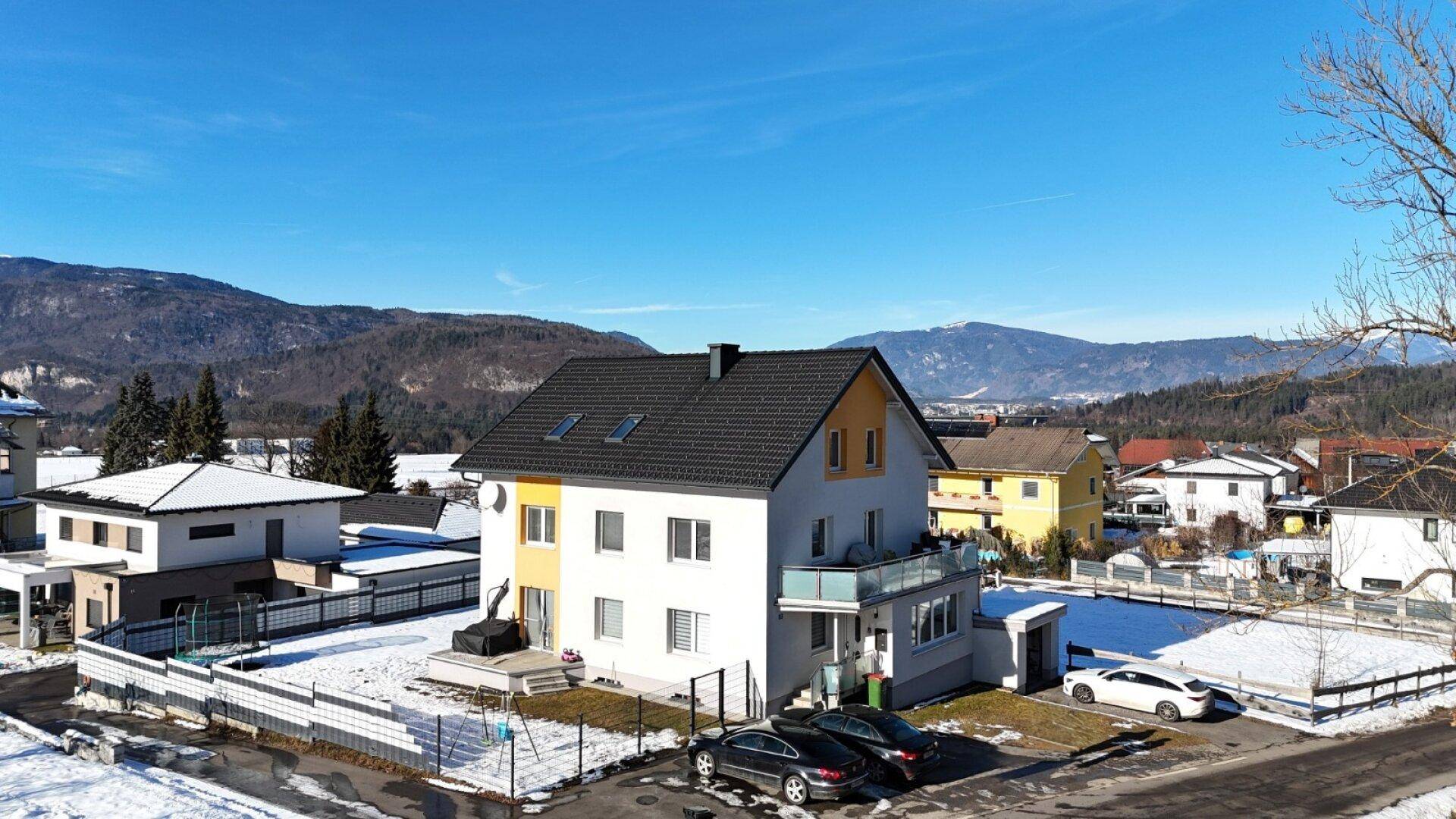 Modernes Einfamilienhaus mit Balkon, Parkplätzen und Bergblick in winterlicher Umgebung.