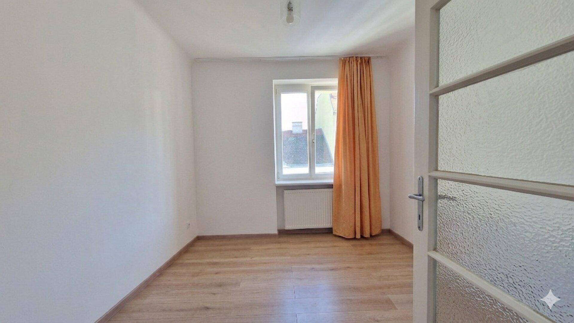 Leeres Zimmer mit Laminatboden, weiß gestrichenen Wänden und einem Fenster mit Vorhang.