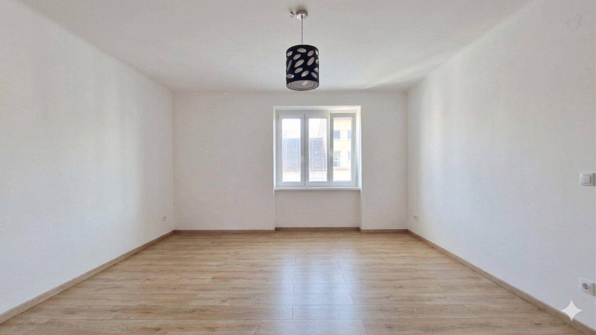 Helles, leeres Zimmer mit Laminatboden, weißen Wänden und einer modernen Deckenleuchte.
