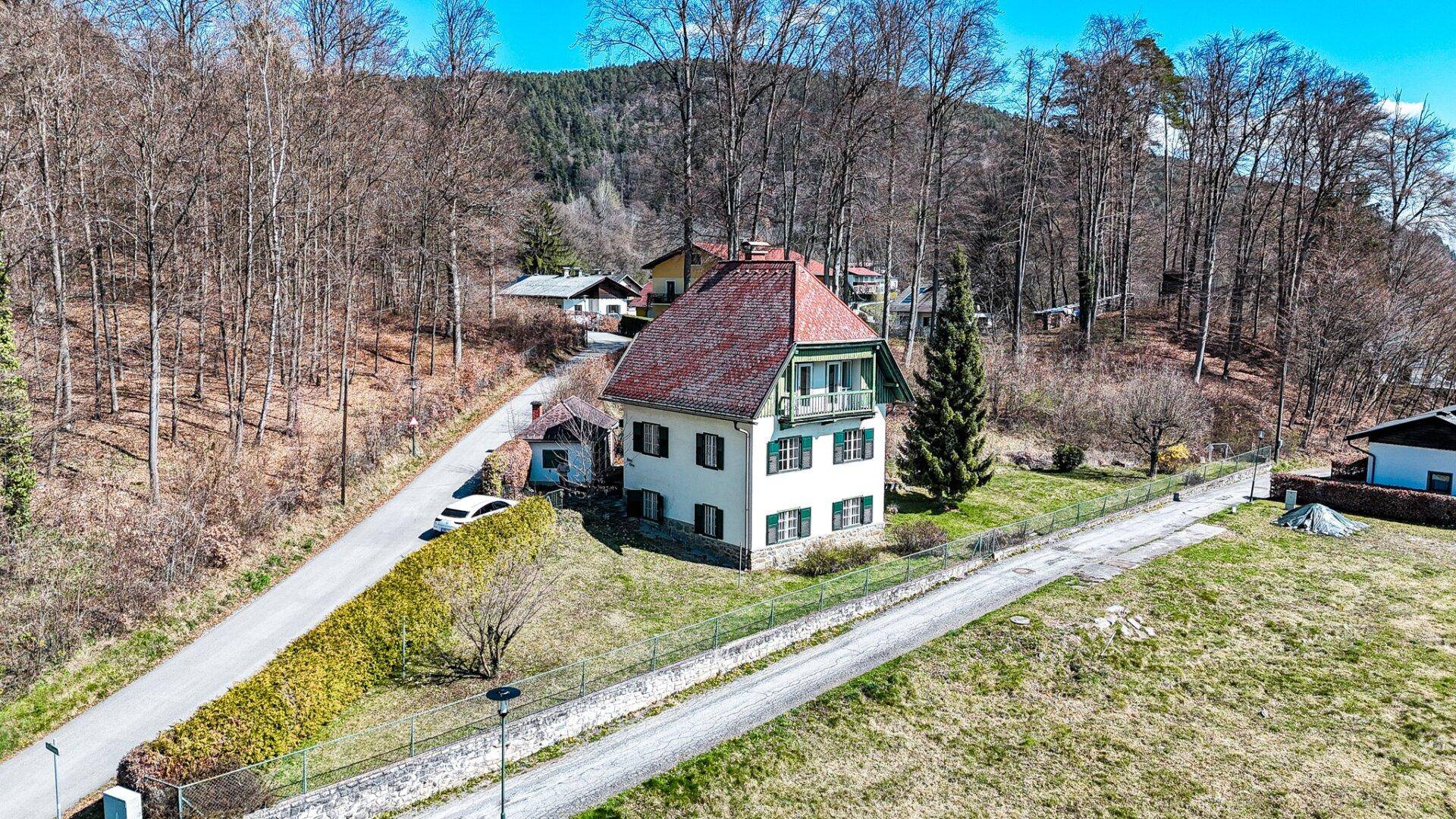 Luftaufnahme eines traditionellen Hauses mit rotem Dach und grünen Fensterläden, umgeben von Bäumen und einer Straße.