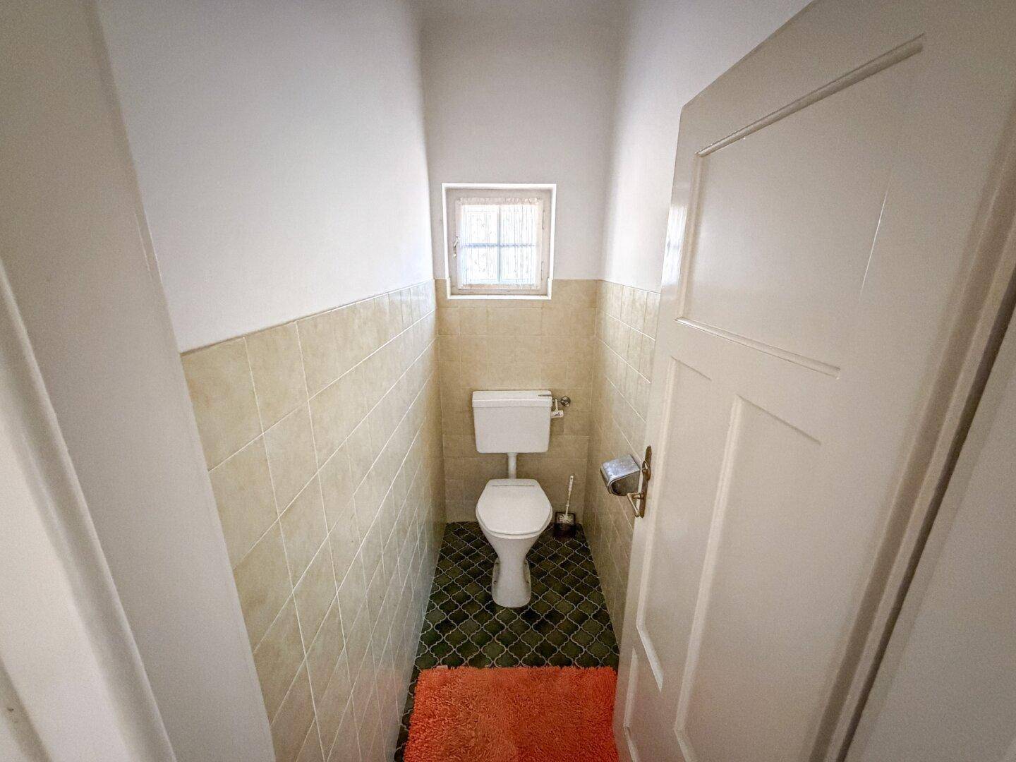 Kleines Gäste-WC mit Fenster, Fliesenboden und einfacher Toilette in hellem Design.