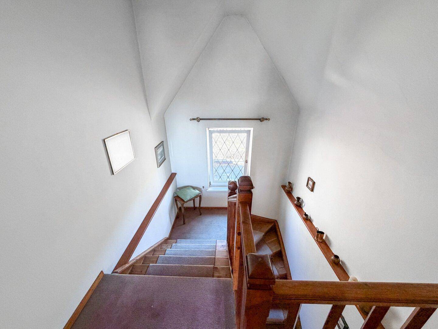 Holztreppe führt in das obere Stockwerk, beleuchtet durch ein kleines Fenster im Dachbereich.