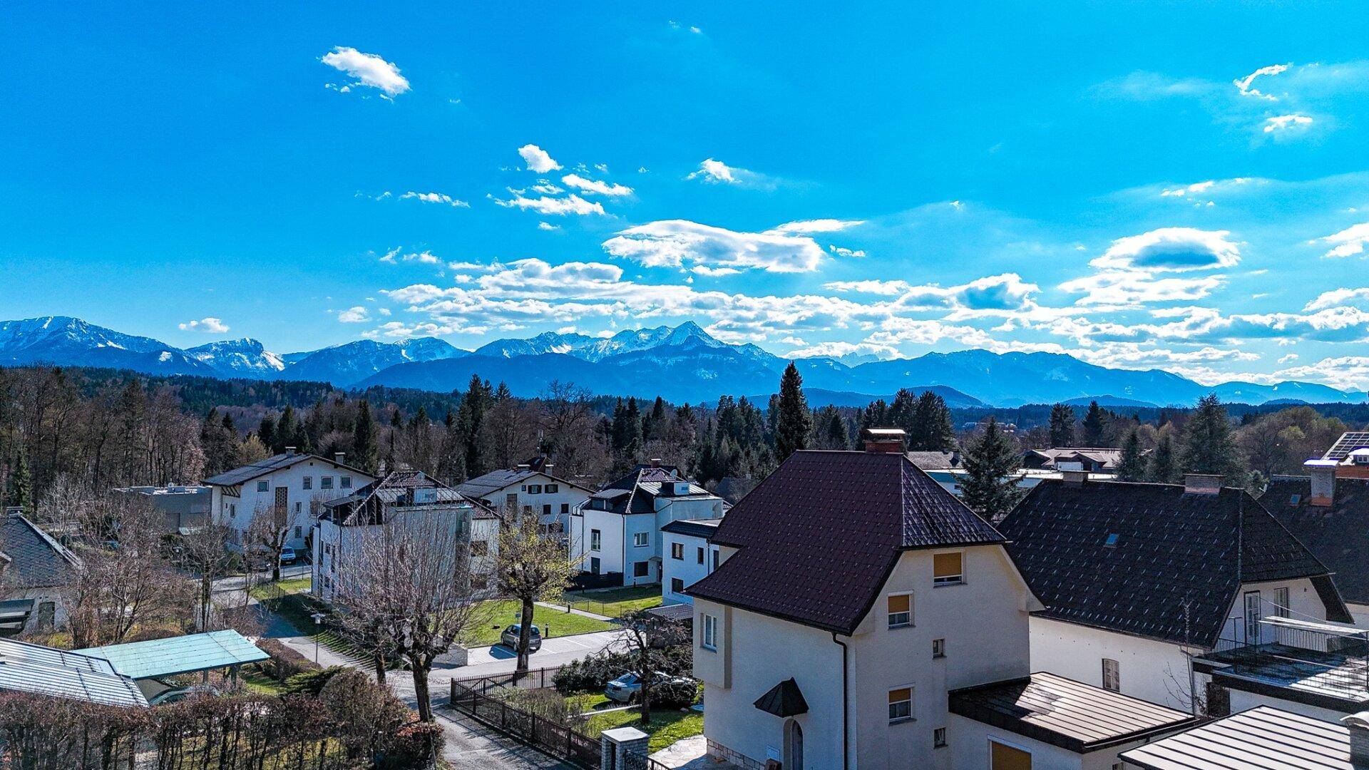 Panoramablick über die Wohngegend mit Häusern und schneebedeckten Bergen im Hintergrund unter blauem Himmel.
