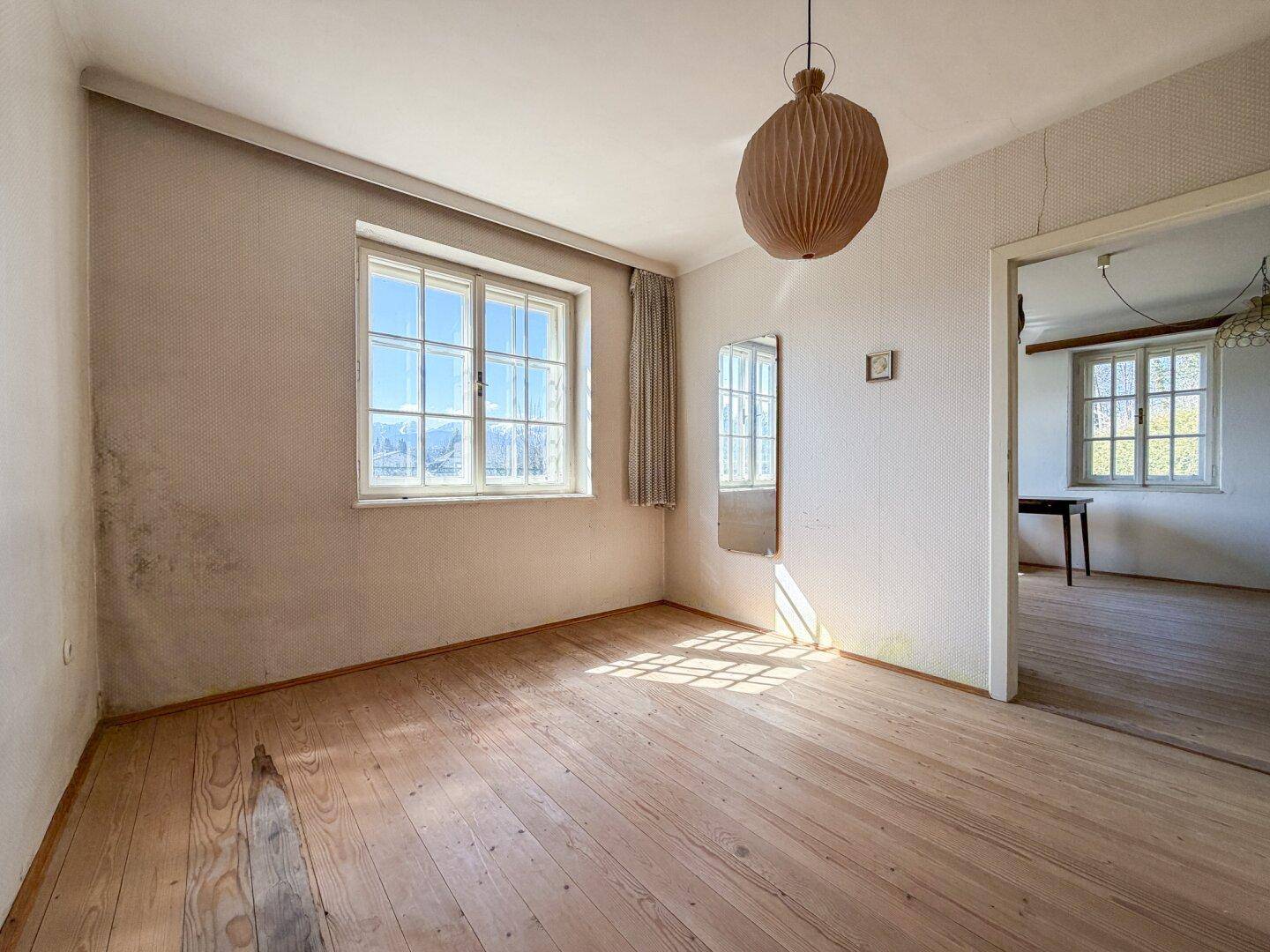 Geräumiges Zimmer mit Holzdielenboden, hellen Wänden und viel Tageslicht durch große Fenster.