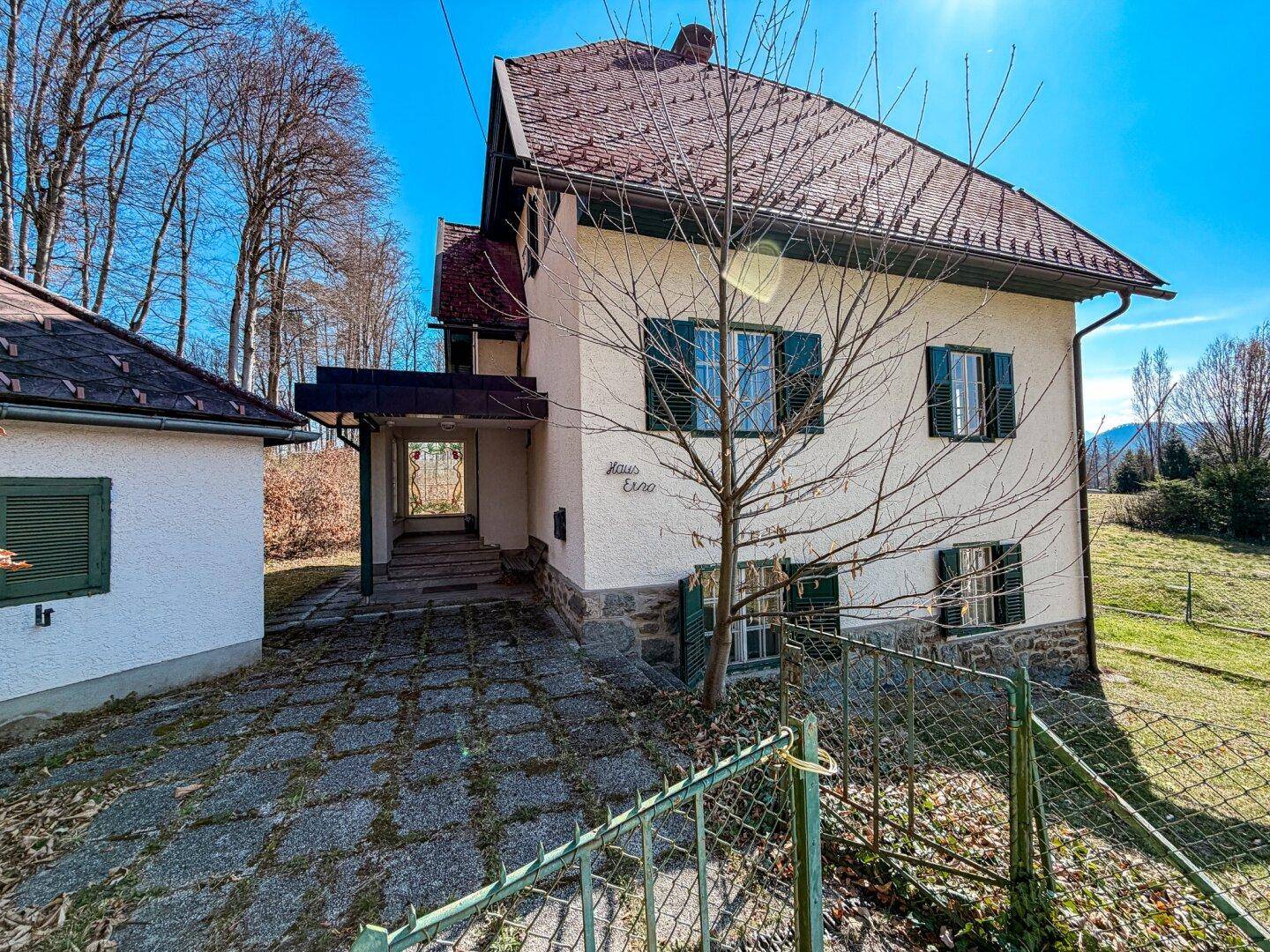 Seitenansicht des Hauses mit gepflastertem Vorplatz und angrenzendem Nebengebäude unter blauem Himmel.