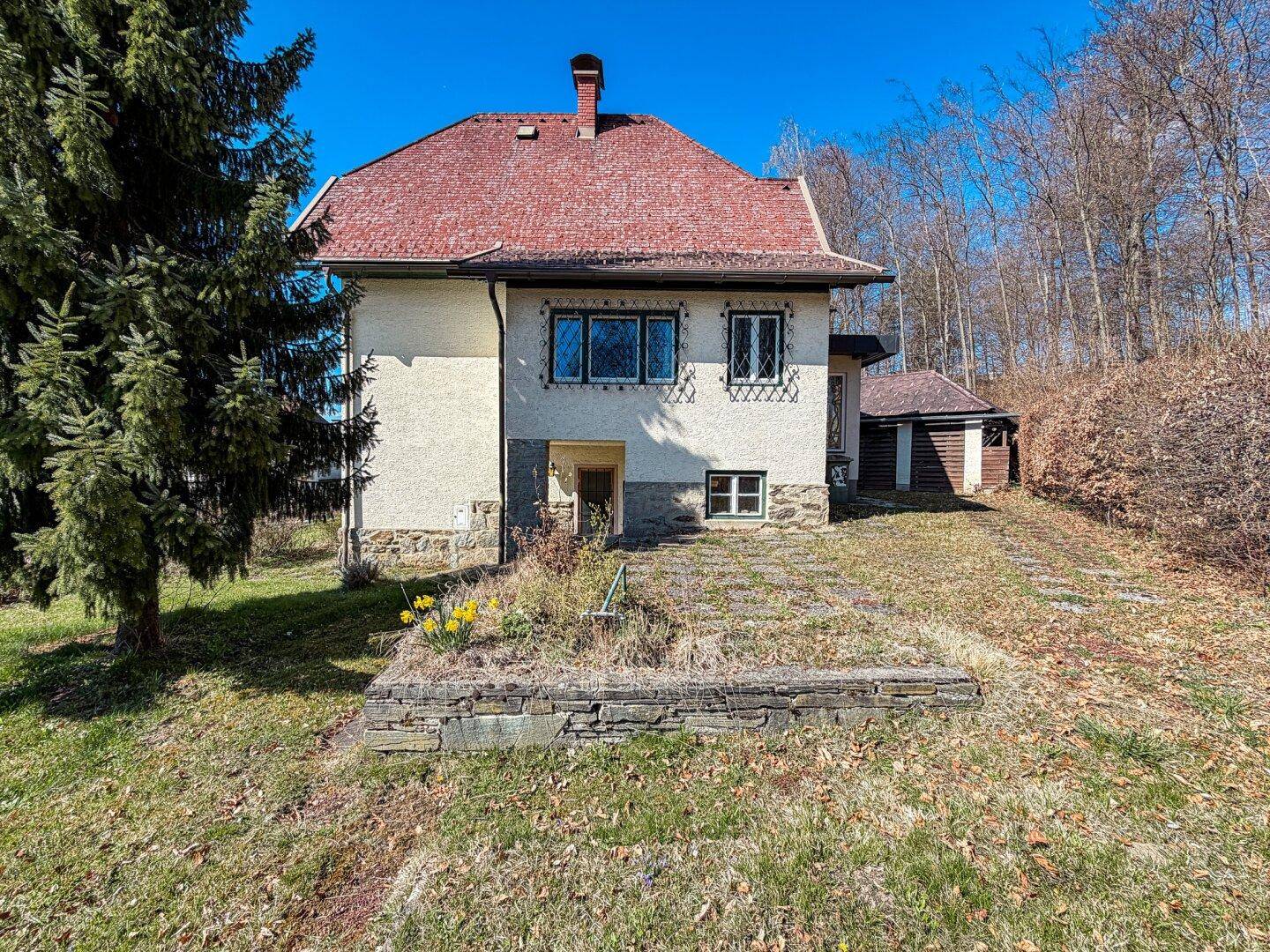 Rückansicht des Hauses mit Gartenbereich, Blumenbeeten und altem Baumbestand in der Sonne.