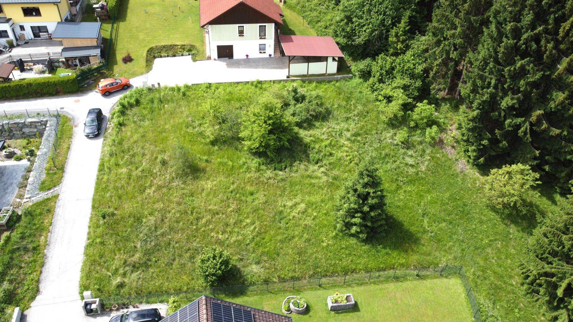 Übersicht aus der Vogelperspektive eines Baugrundstücks mit angrenzendem Wald und Wohnhaus.