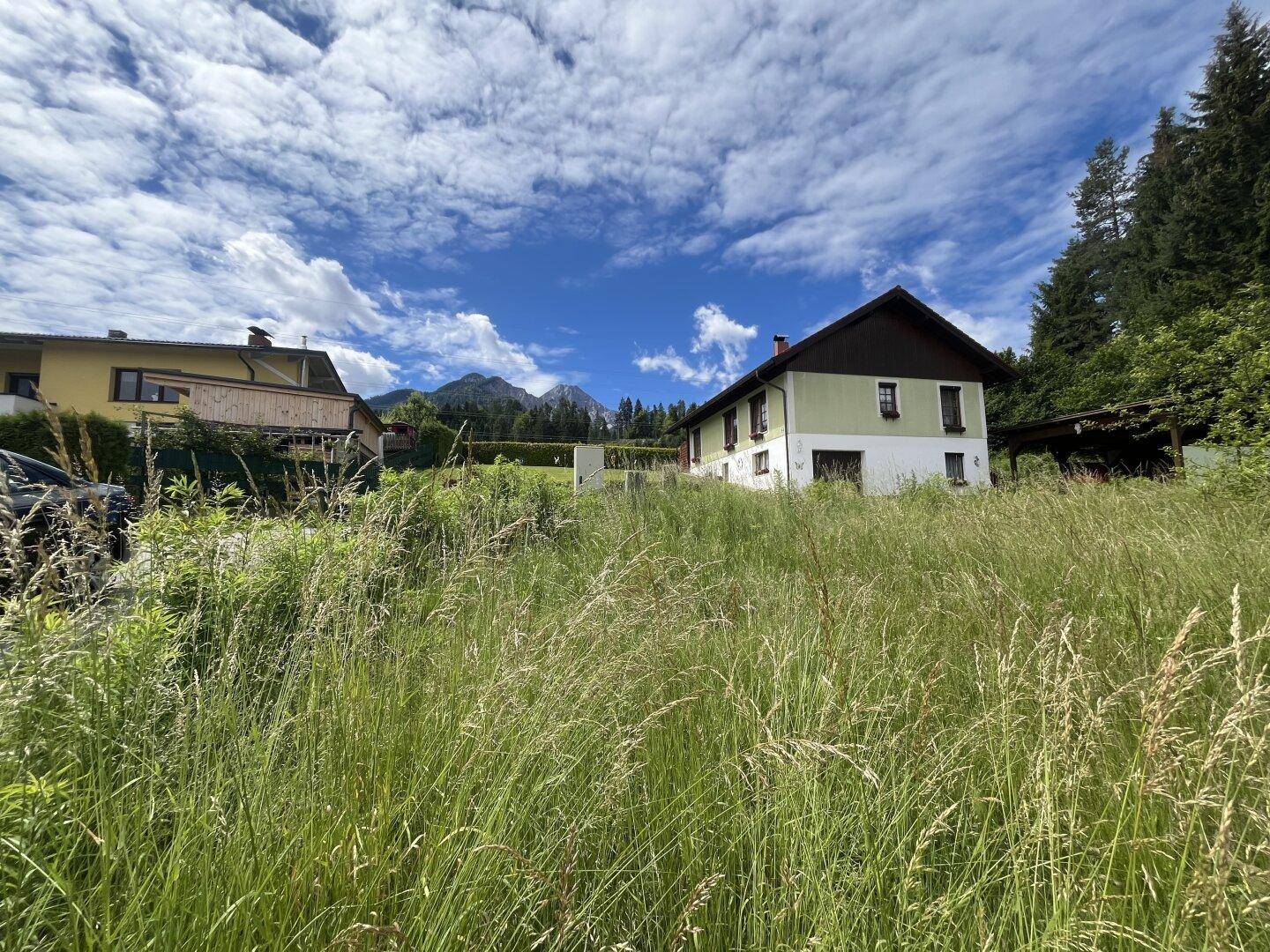 Blick über eine grüne Wiese auf ein Wohnhaus mit Bergen im Hintergrund unter blauem Himmel.
