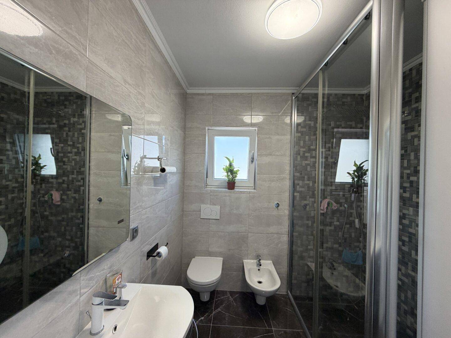 Modernes Badezimmer mit Dusche, WC, Bidet und einem Fenster für natürliches Licht und Belüftung.