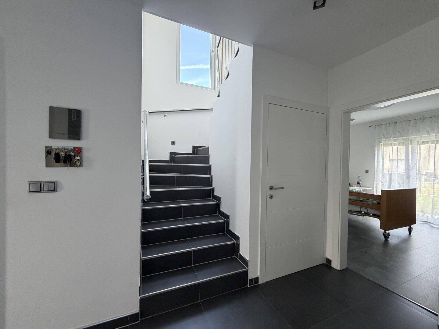 Heller Eingangsbereich mit moderner Treppe und dunklen Fliesen, die zu den oberen Etagen führt.