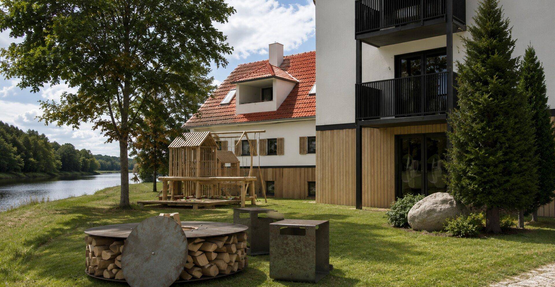Hausfassade mit Balkonen, umgeben von Grünflächen, einem Spielplatz und einem Feuerplatz am Seeufer.