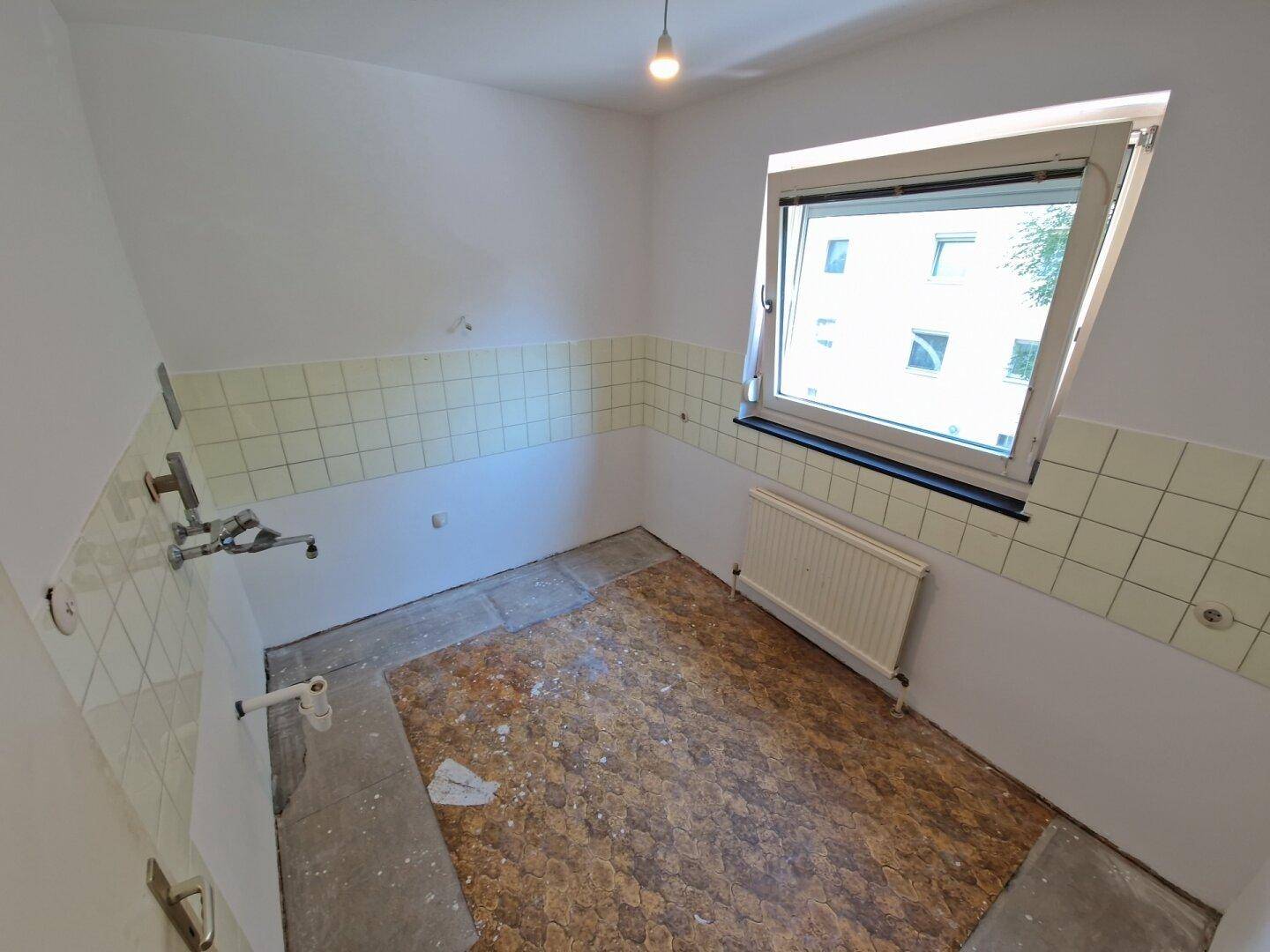 Leere Küche mit Fenster, Heizkörper und teilweise gefliesten Wänden, bereit für die Renovierung.