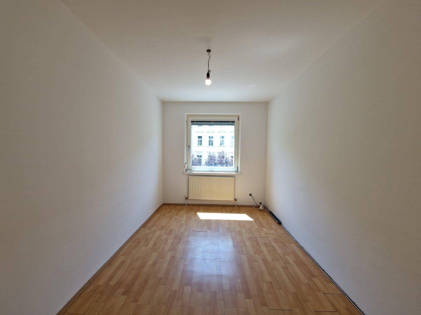 Schmaler, heller Raum mit Laminatboden, Fenster und Heizkörper, gut geeignet als Schlaf- oder Arbeitszimmer.