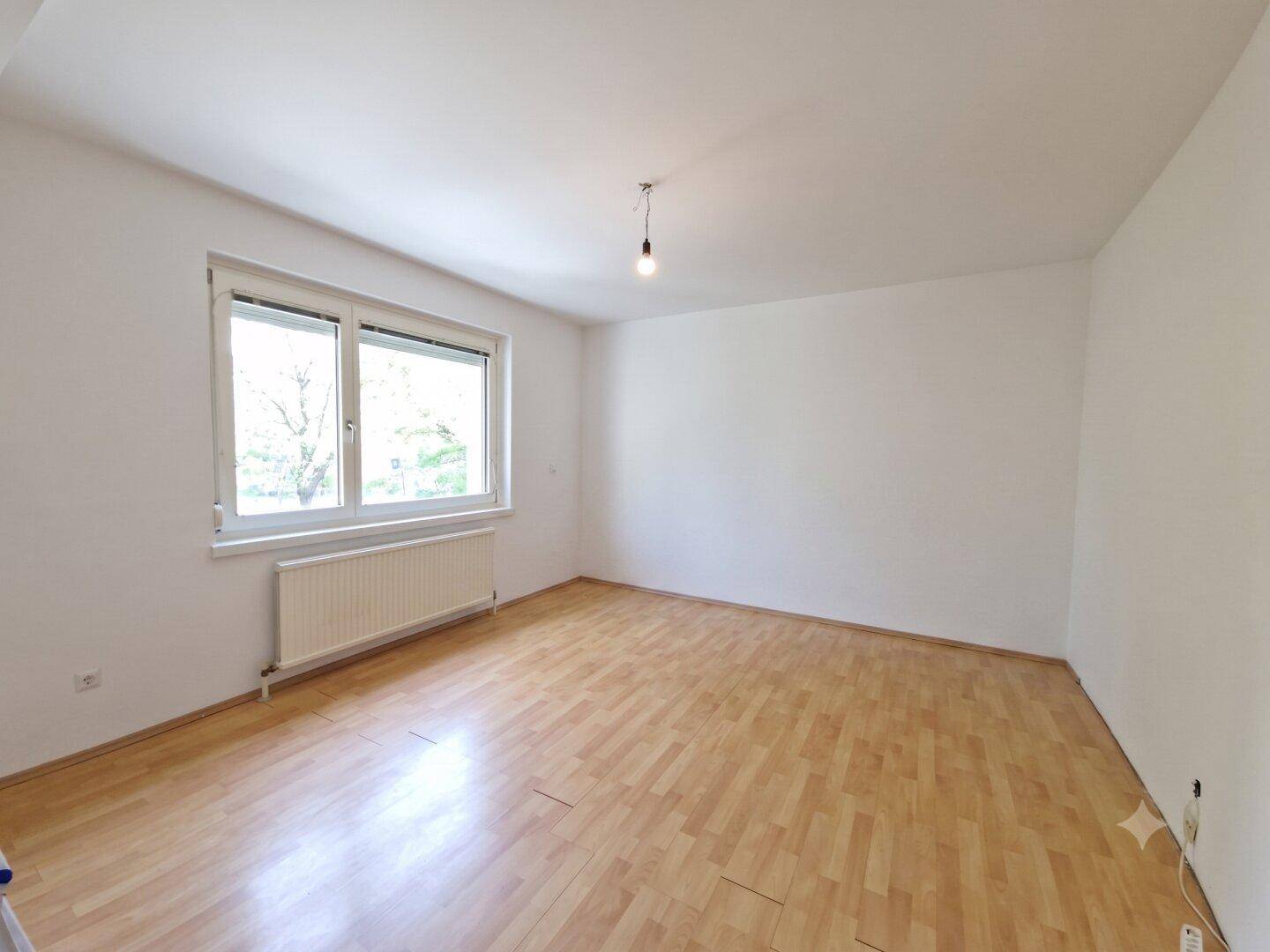 Heller, leerer Raum mit Laminatboden, großem Fenster und Heizkörper, ideal als Wohn- oder Schlafzimmer.
