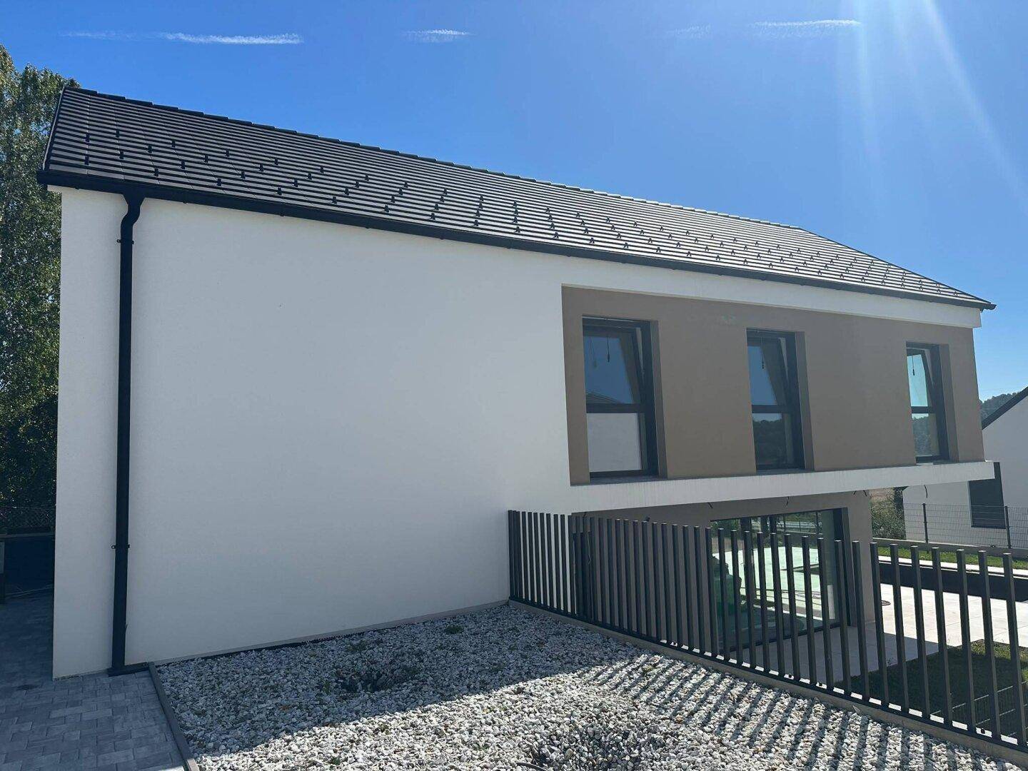 Seitenansicht des modernen Hauses mit heller Fassade, dunklem Dach und einem Kiesbeet.