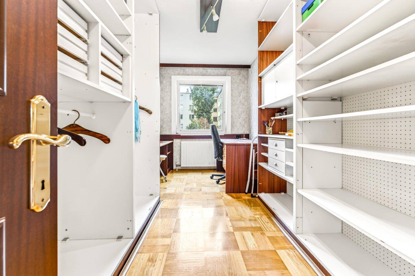 Praktischer begehbarer Kleiderschrank führt zu einem hellen Arbeitszimmer mit Fenster.