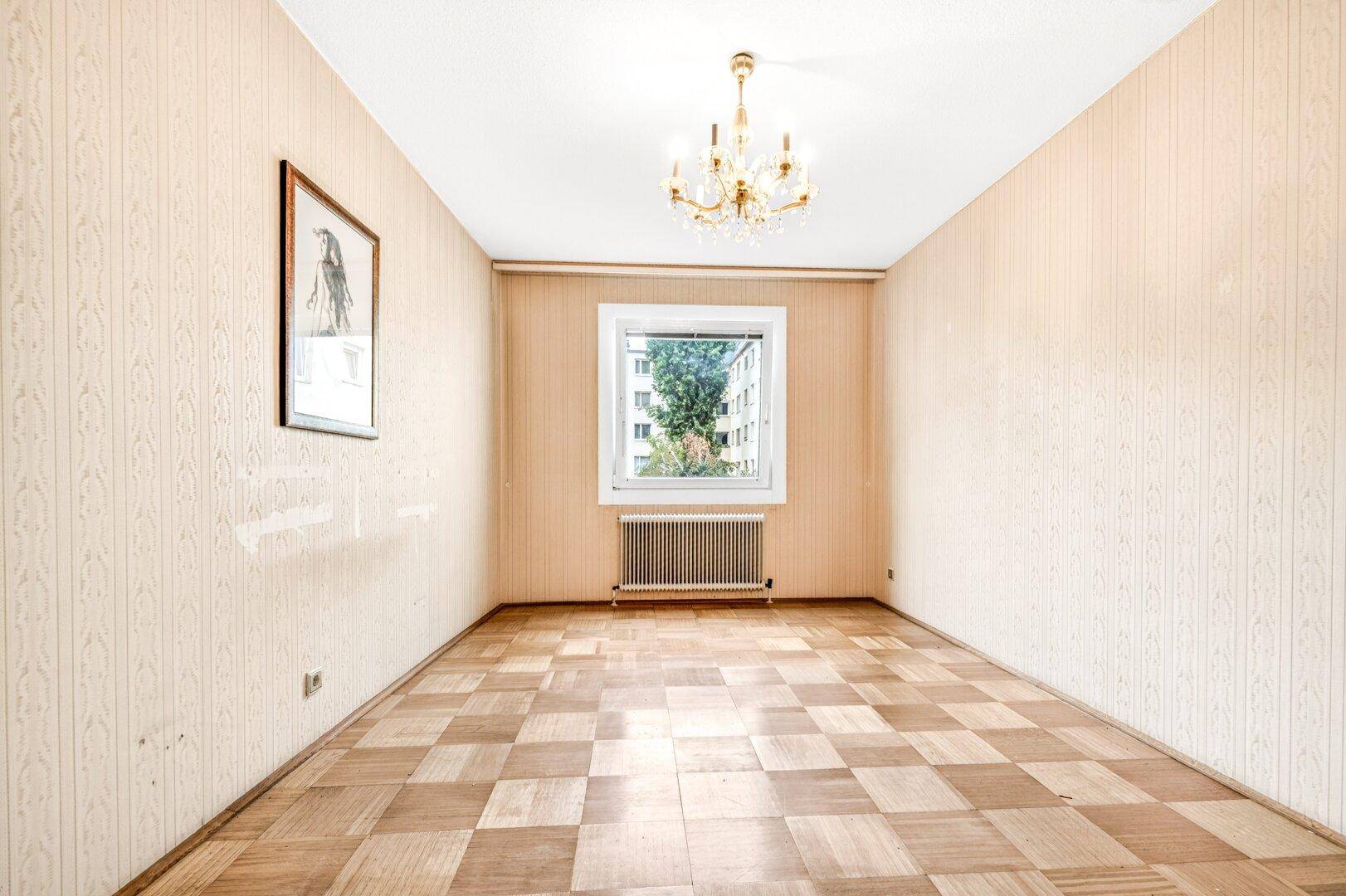 Leeres Zimmer mit Parkettboden, gemusterter Tapete und einem Fenster mit Blick ins Grüne.
