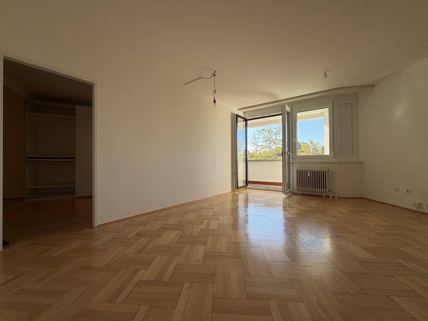 Geräumiges Wohnzimmer mit Parkettboden und offener Balkontür, die viel Licht hereinlässt.