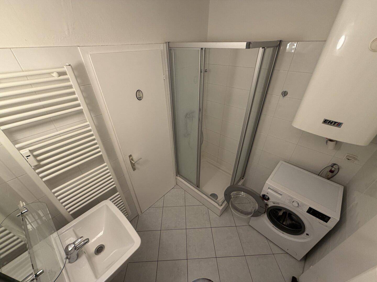 Kompaktes Badezimmer mit Dusche, Waschbecken, Waschmaschine und beheiztem Handtuchhalter.