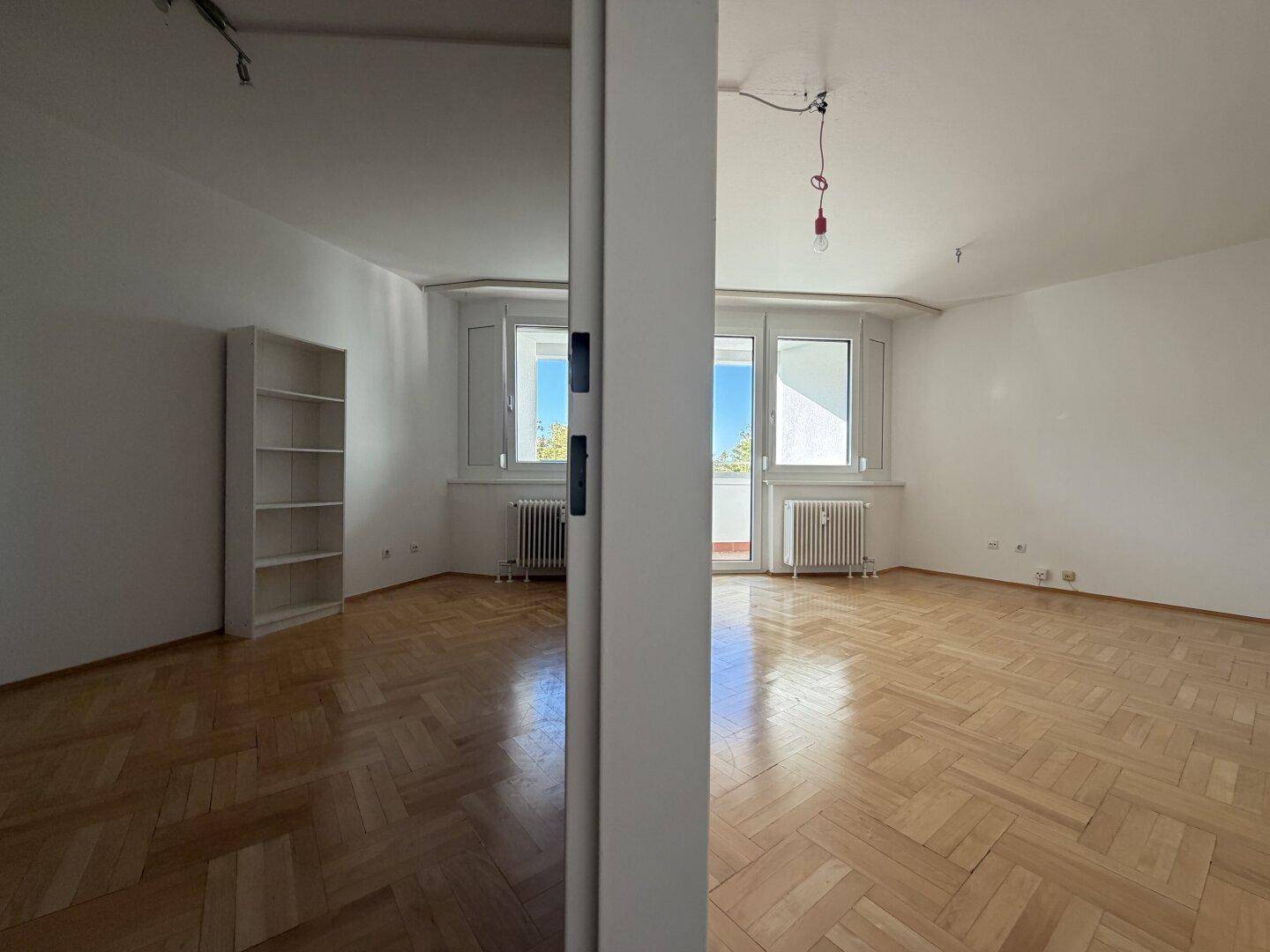 Großer Wohnbereich mit Parkettboden, Fensterfront und einem schlichten Bücherregal.