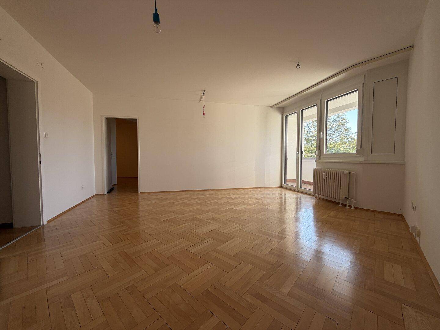 Heller, leerer Wohnraum mit Fischgrät-Parkettboden und großen Fenstern zum Balkon.