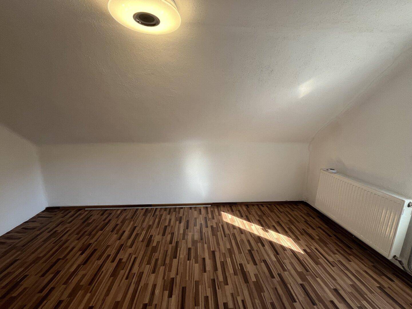Helles Zimmer im Dachgeschoss mit Laminatboden, Heizkörper und schräger Decke.