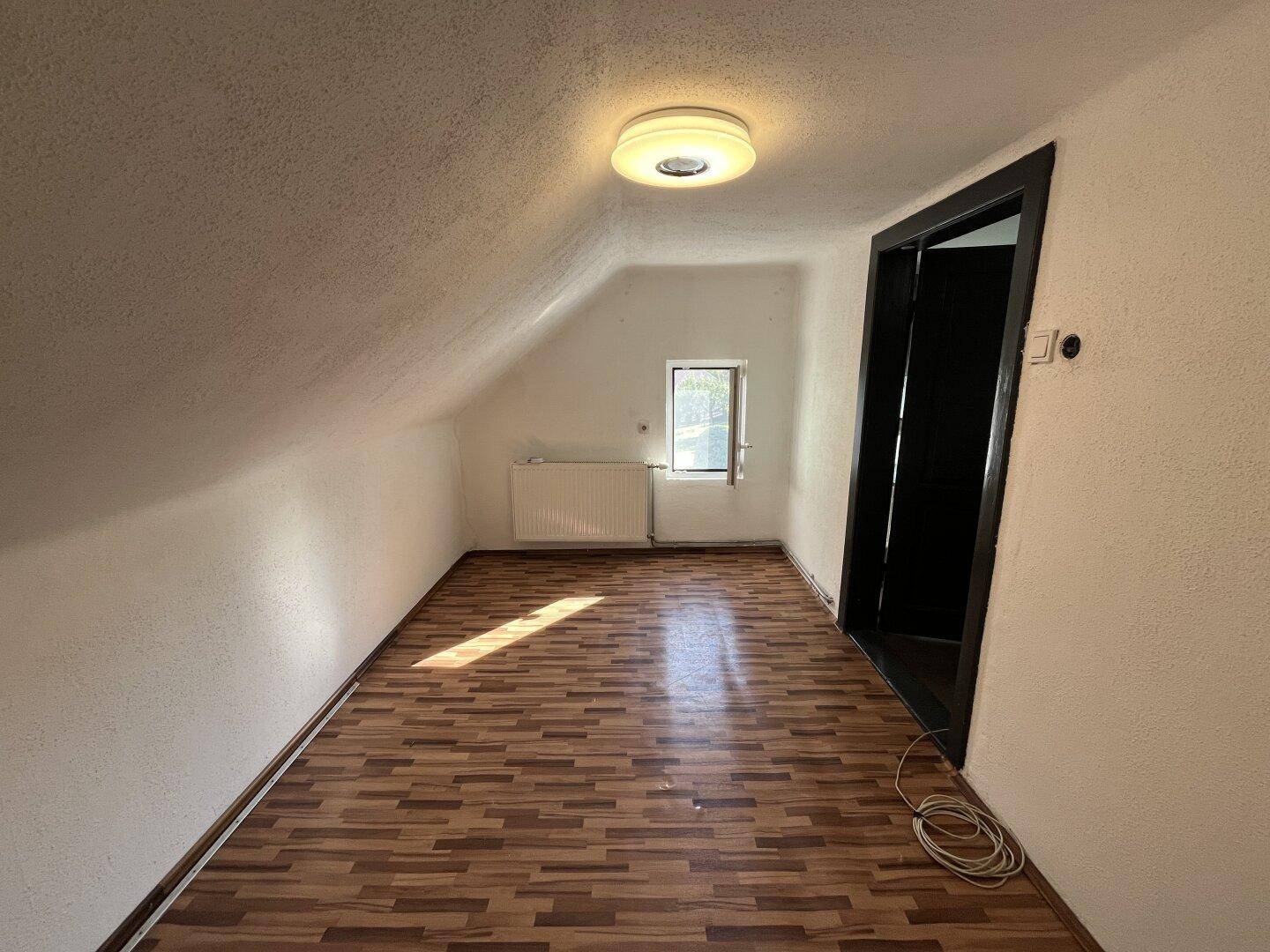 Leeres Zimmer im Dachgeschoss mit Laminatboden, kleinem Fenster und schrägen Wänden.