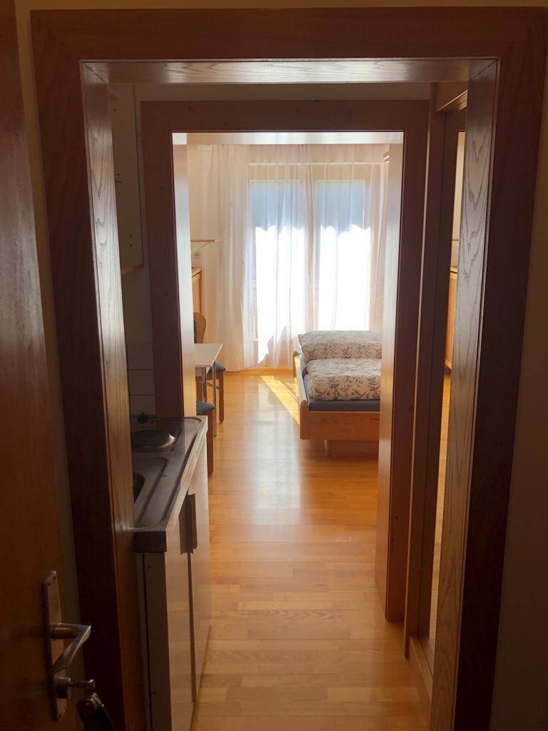Blick durch einen Türrahmen in einen Raum mit Holzboden und Bett, links eine kleine Küchenzeile.