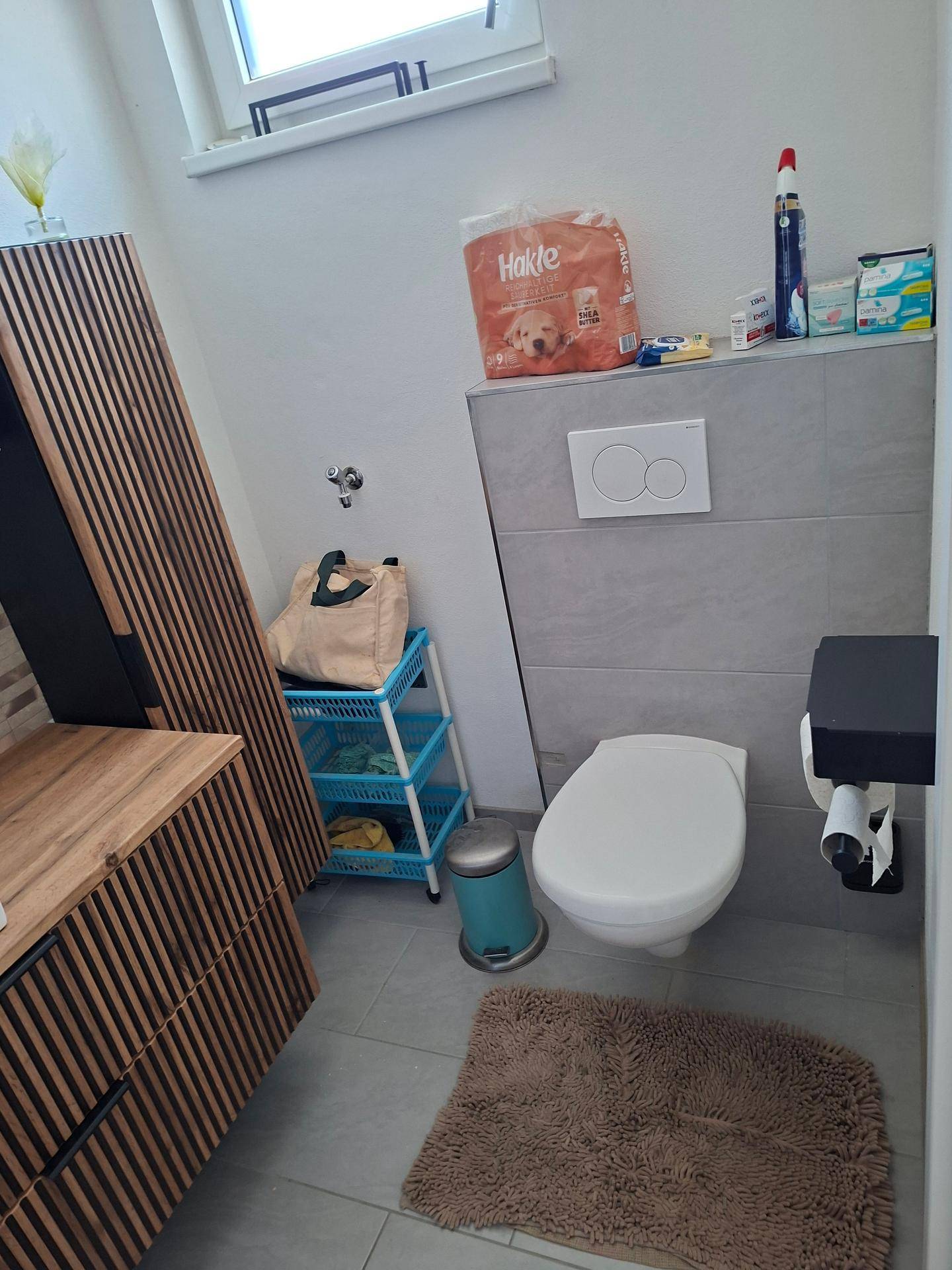 Badezimmer mit wandhängendem WC, Fliesenboden und einem Fenster für Tageslicht.