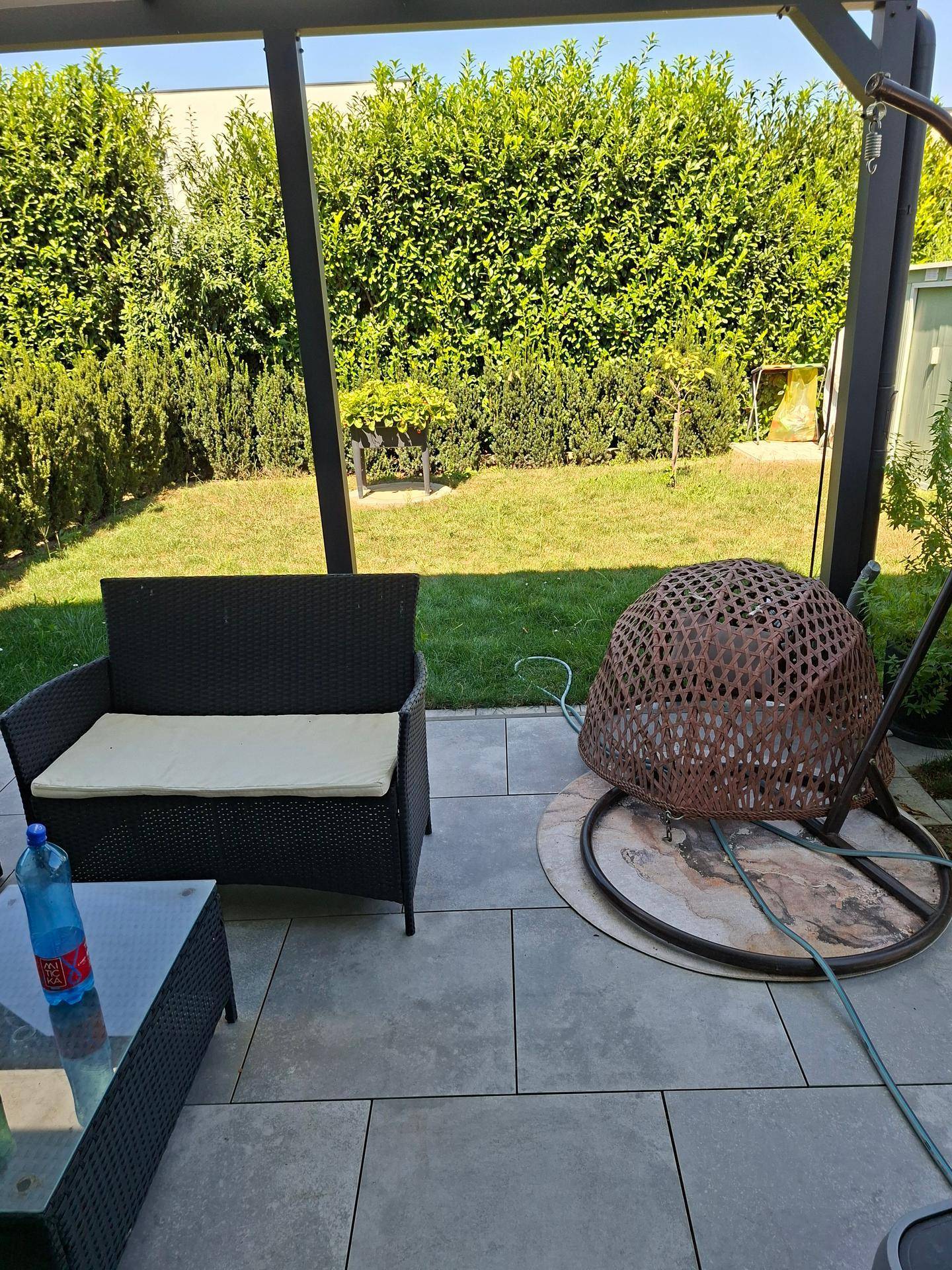 Sonniger Terrassenbereich mit Gartenmöbeln, umgeben von einer grünen Hecke und Rasenfläche.