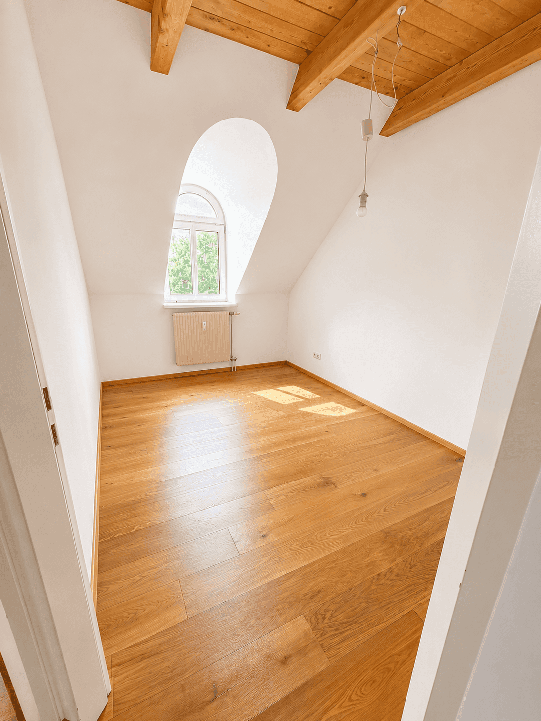 Leeres Schlafzimmer im Dachgeschoss mit Holzboden, sichtbaren Balken und Rundbogenfenster.