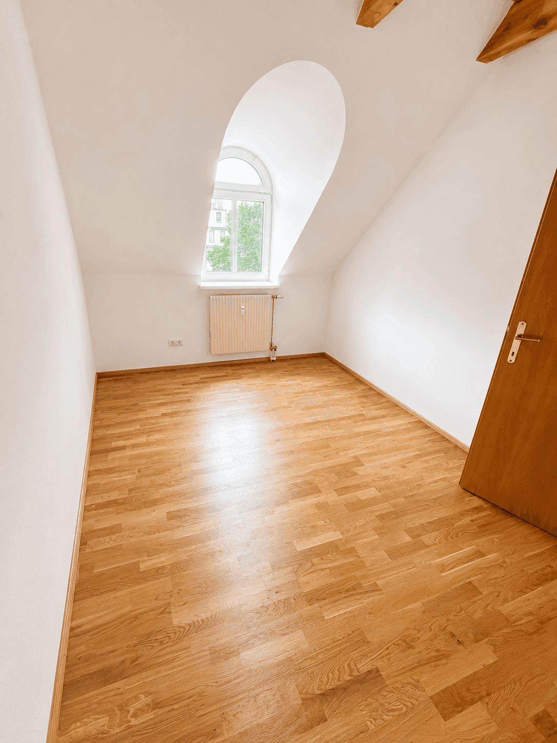 Helles Schlafzimmer im Dachgeschoss mit Holzboden, Heizkörper und Rundbogenfenster.