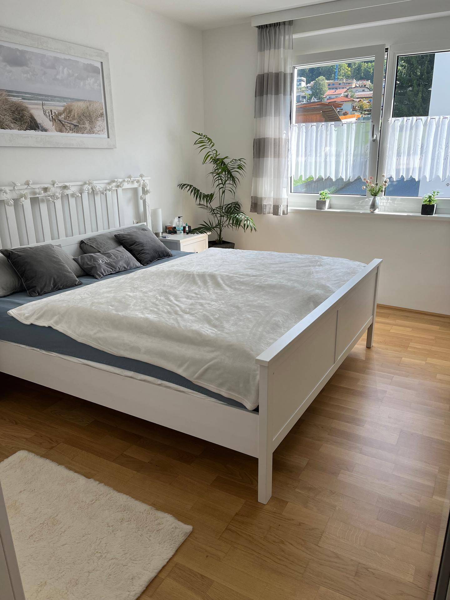Helles Schlafzimmer mit weißem Bettrahmen, Holzboden und Blick ins Grüne.