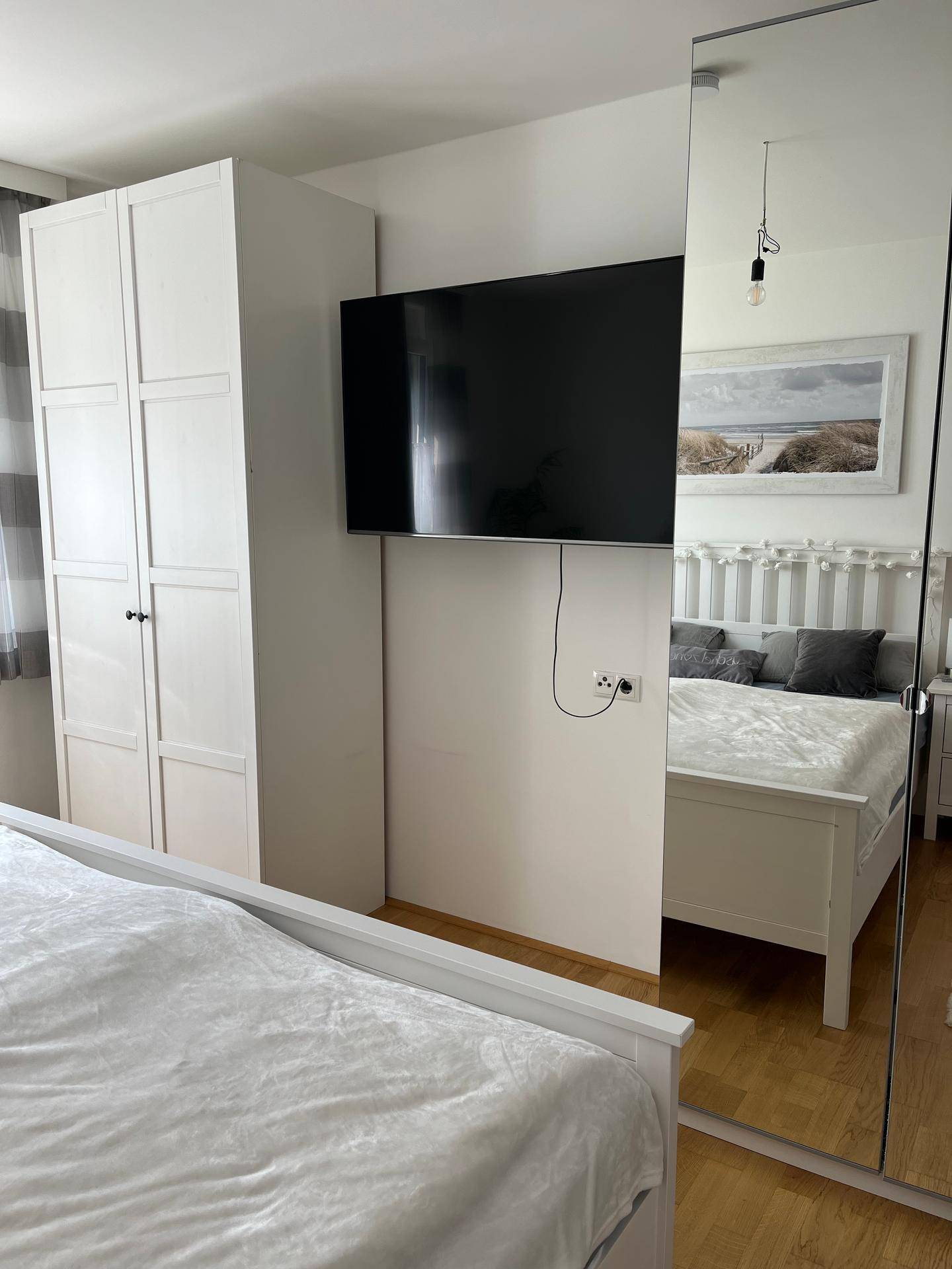 Schlafzimmer mit Wandfernseher, weißem Kleiderschrank und großem Spiegel.