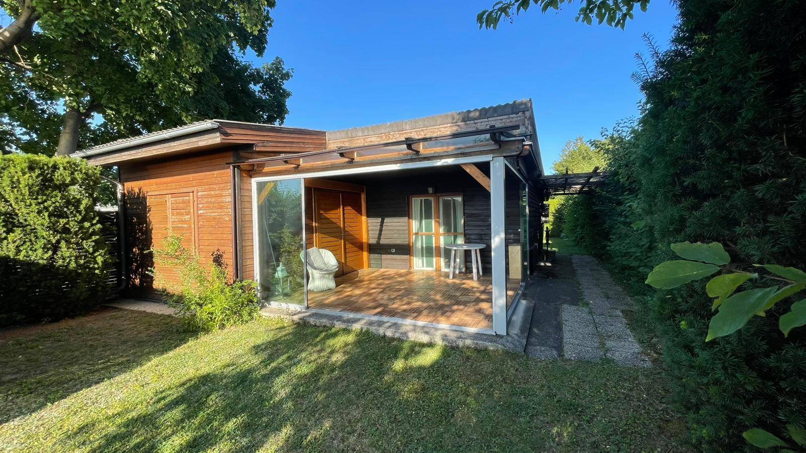 Holzhaus mit überdachter Terrasse und Glasfront, umgeben von gepflegtem Rasen und Hecken.