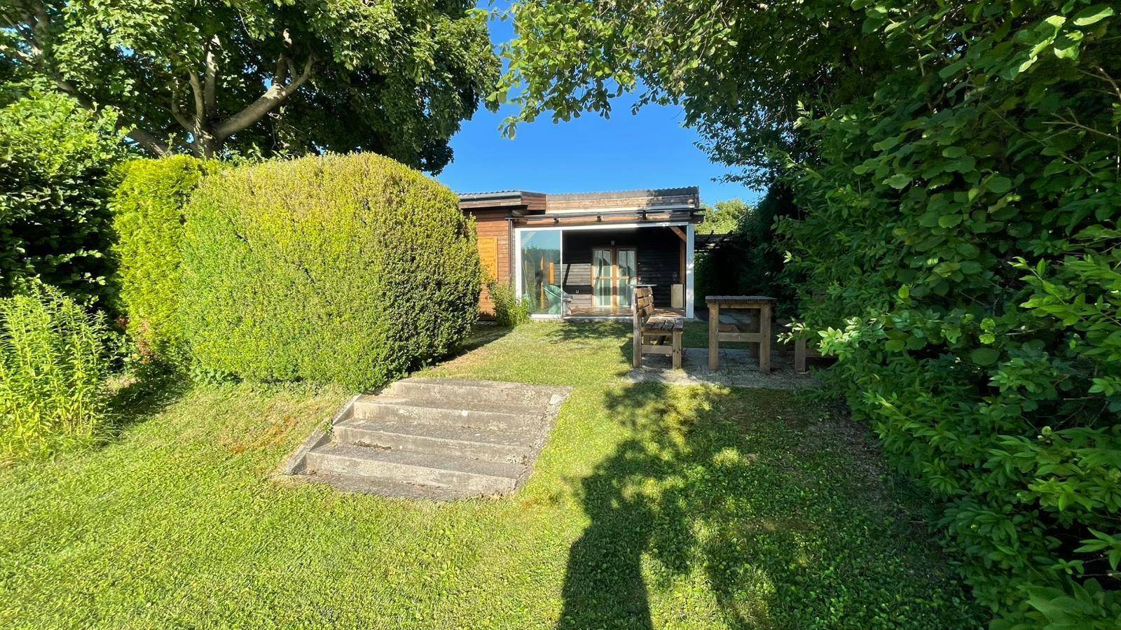 Kleines Holzhaus umgeben von üppigem Grün mit Treppe und Sitzgelegenheit im Garten.
