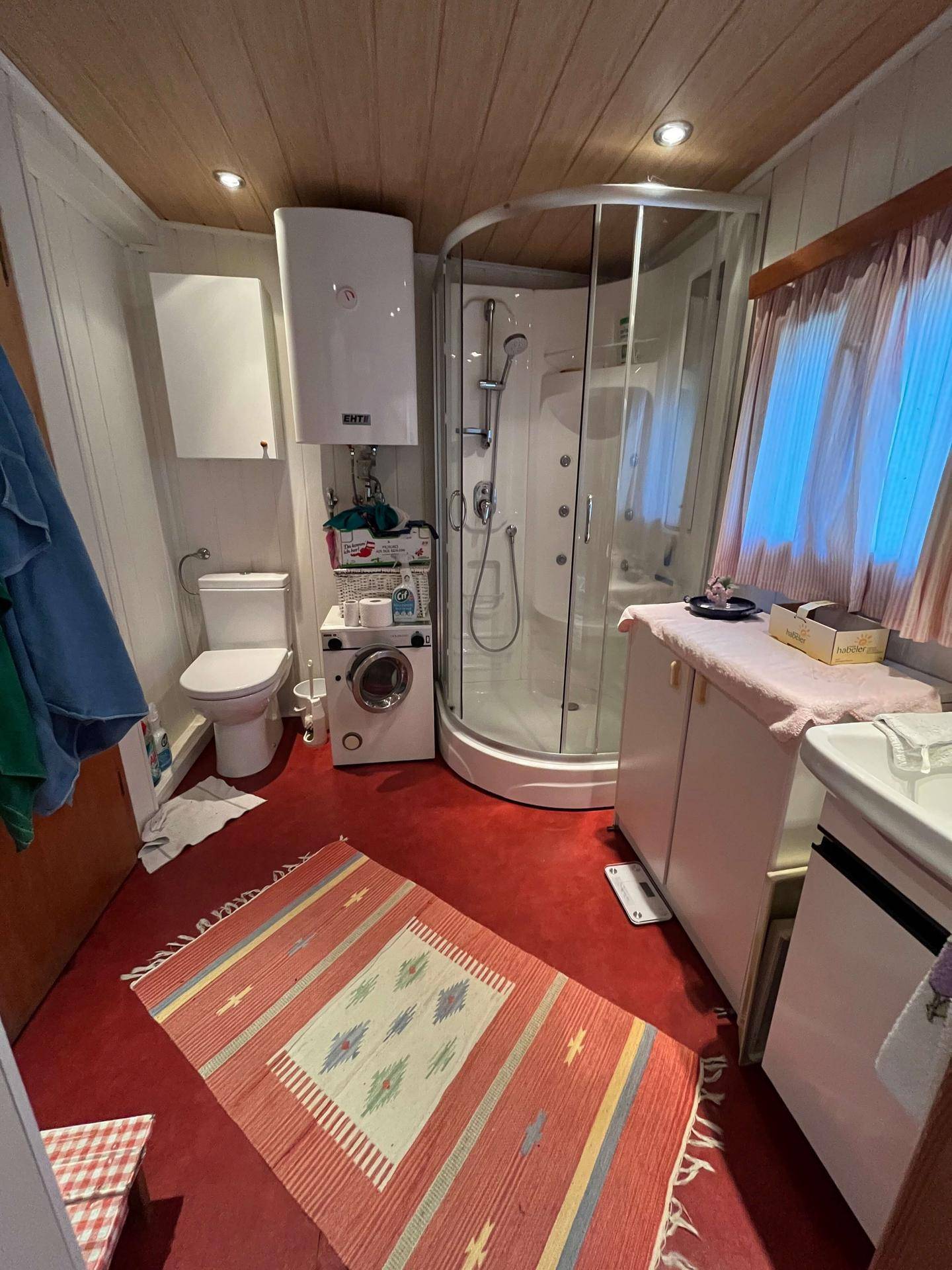 Kompaktes Badezimmer mit Duschkabine, Toilette, Waschmaschine und rotem Boden.